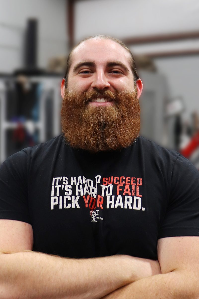 Smiling man with a beard wearing a motivational t-shirt in a gym setting.